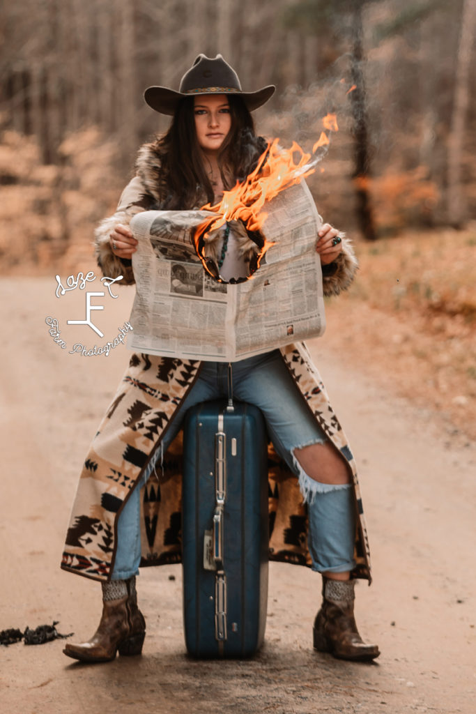 cowgirl sitting on suitcase burning a news paper