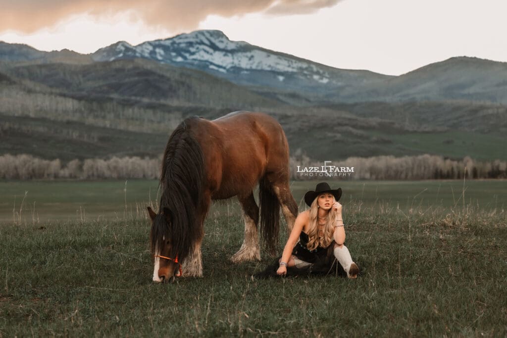 cowgirl in a black dress sitting beside of her horse in a field at sunset with the rocky mountains behind her