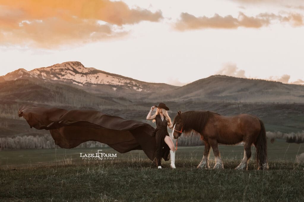 cowgirl in a black flying dress with her horse and cowboy hat at sunset in front of the rocky mountains