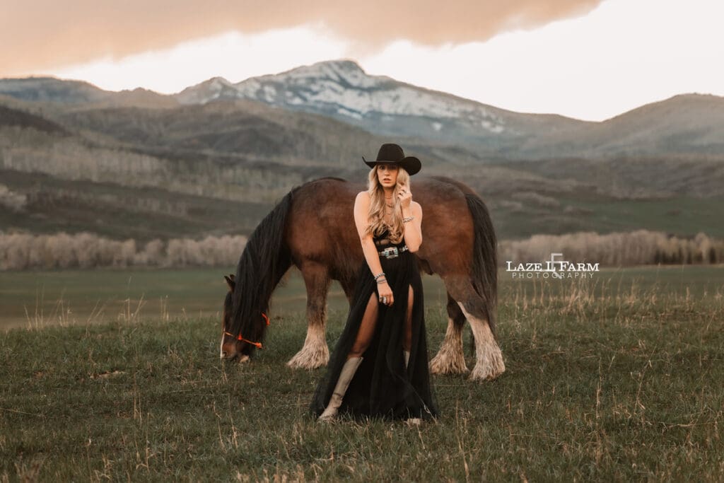 cowgirl wearing black dress with horse and the rocky mountains in the background at sunset