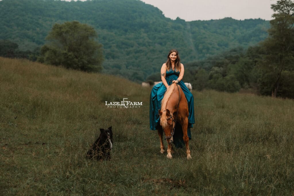 cowgirl sitting on her horse in a field with mountains behind her with her dog