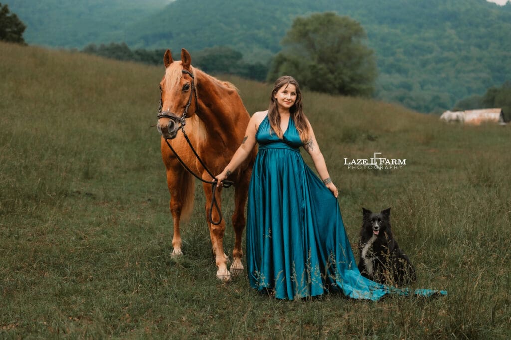 girl standing beside her horse in a field with mountains and her doge beside of her