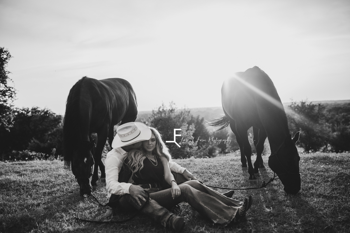 cowbo yand wife sitting on the ground with 2 horses in black and white