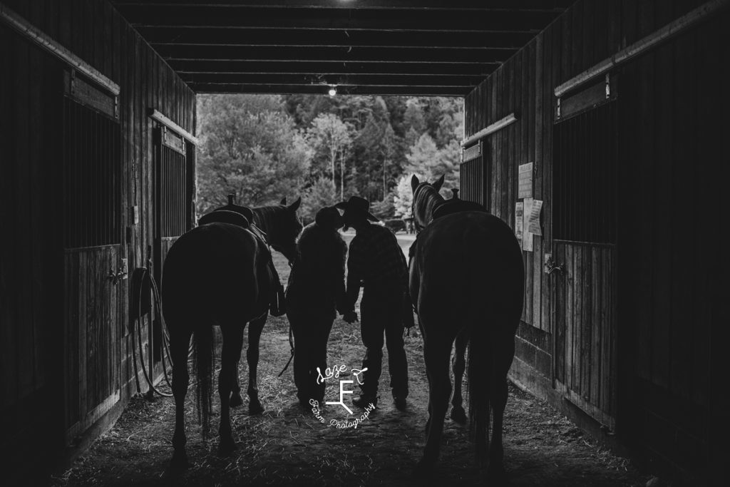 couple with horses kissing in barn aisle in black and white