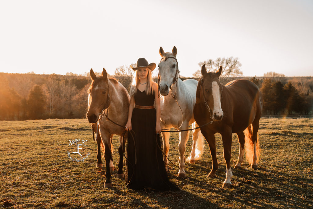 cowgirl in black dress with 3 horses at sunset