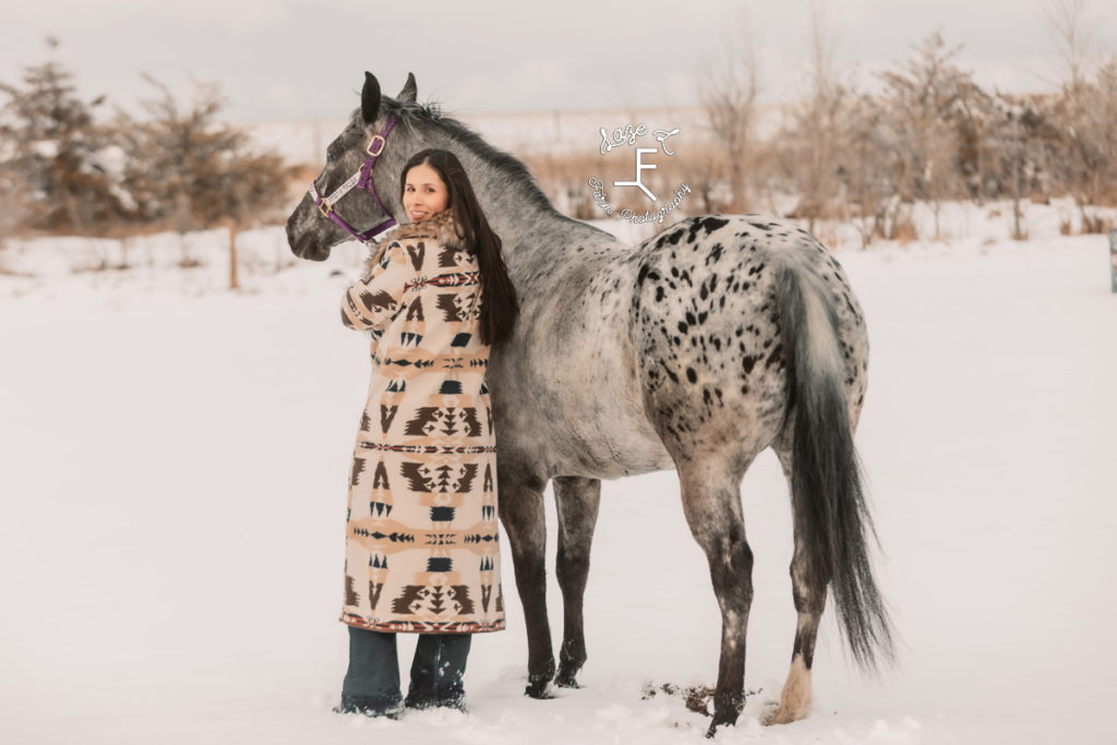 girl in long fur trimmed coat in the snow with appaloosa looking back