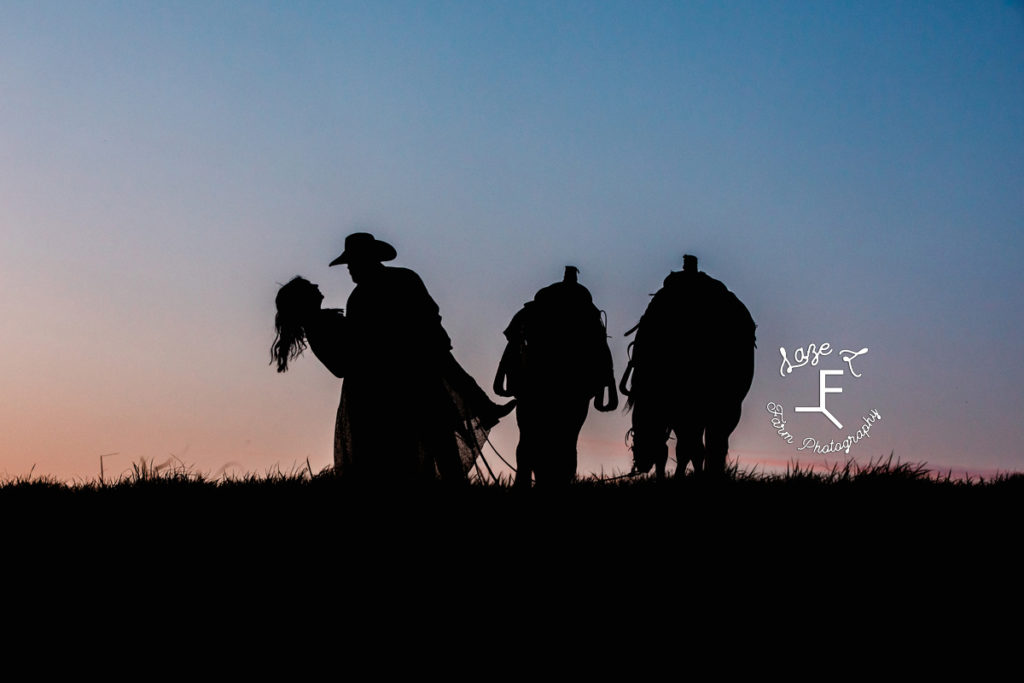 cowboy and wife embracing with 2 horses at sunset silhouette