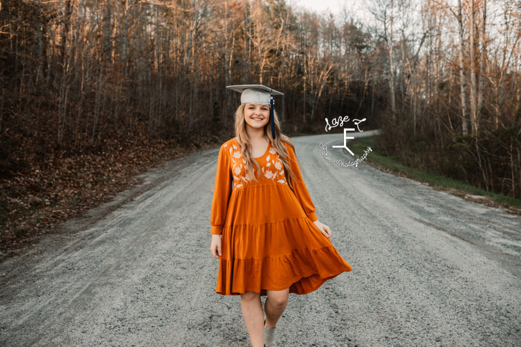 senior girl with cap on in orange dress walking down dirt road