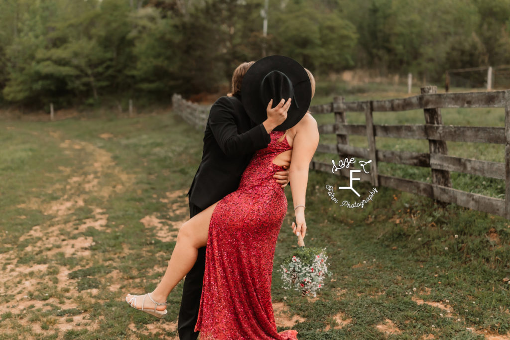 prom couple in red and black kissing behind cowboy hat