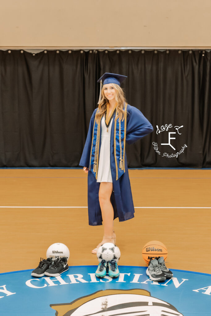 senior girl in cap and gown with 3 sports balls