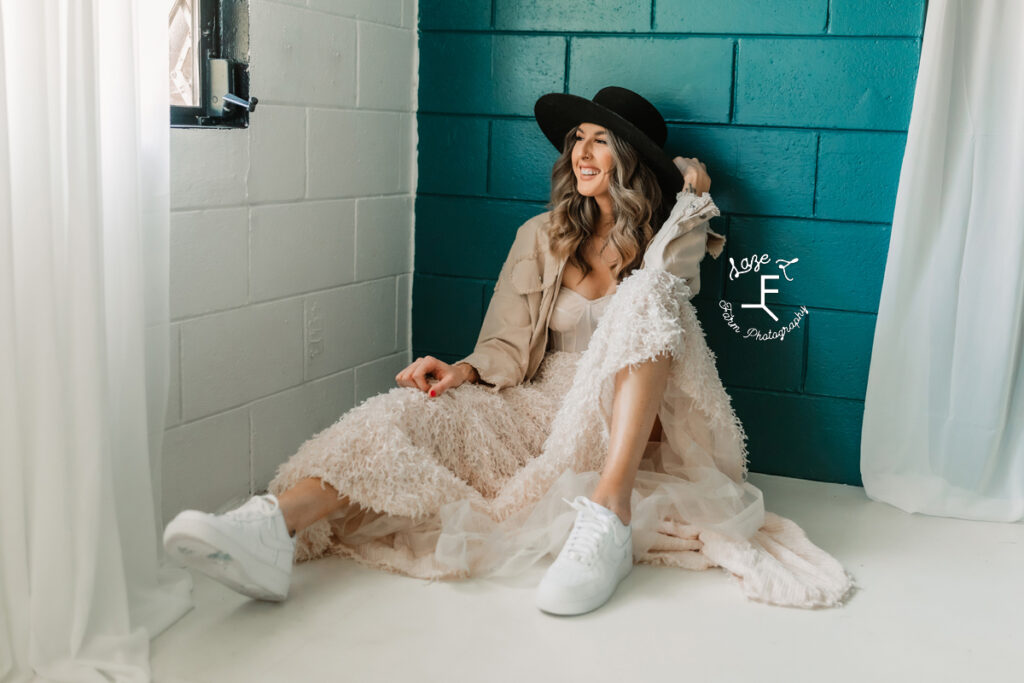 jess sitting against teal wall in white dress and black hat