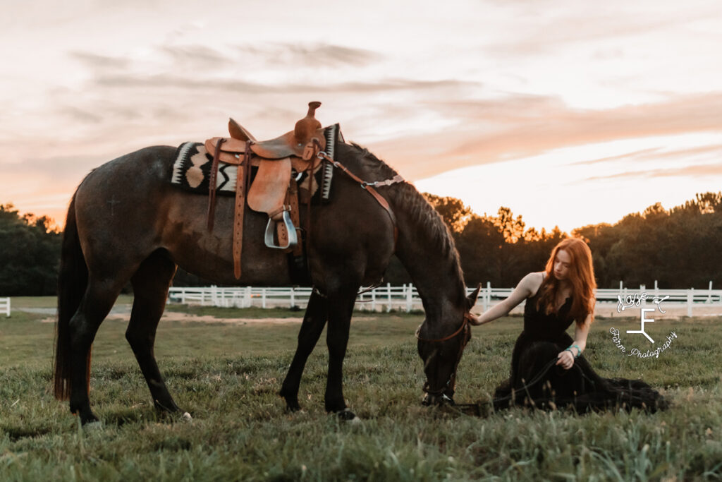cowgirl sitting in grass in black dress with bay roan at sunset
