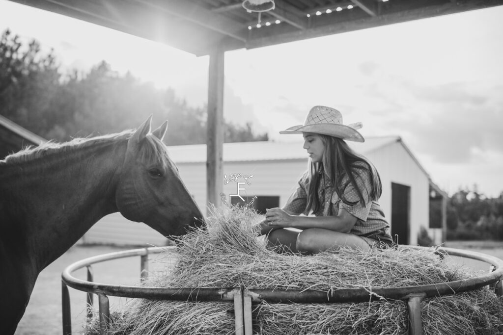 cowgirl sitting on hay petting horse in black and white