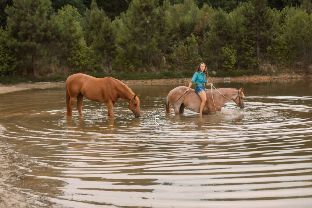 cowgirl riding red roan in to pond while ponying another horse