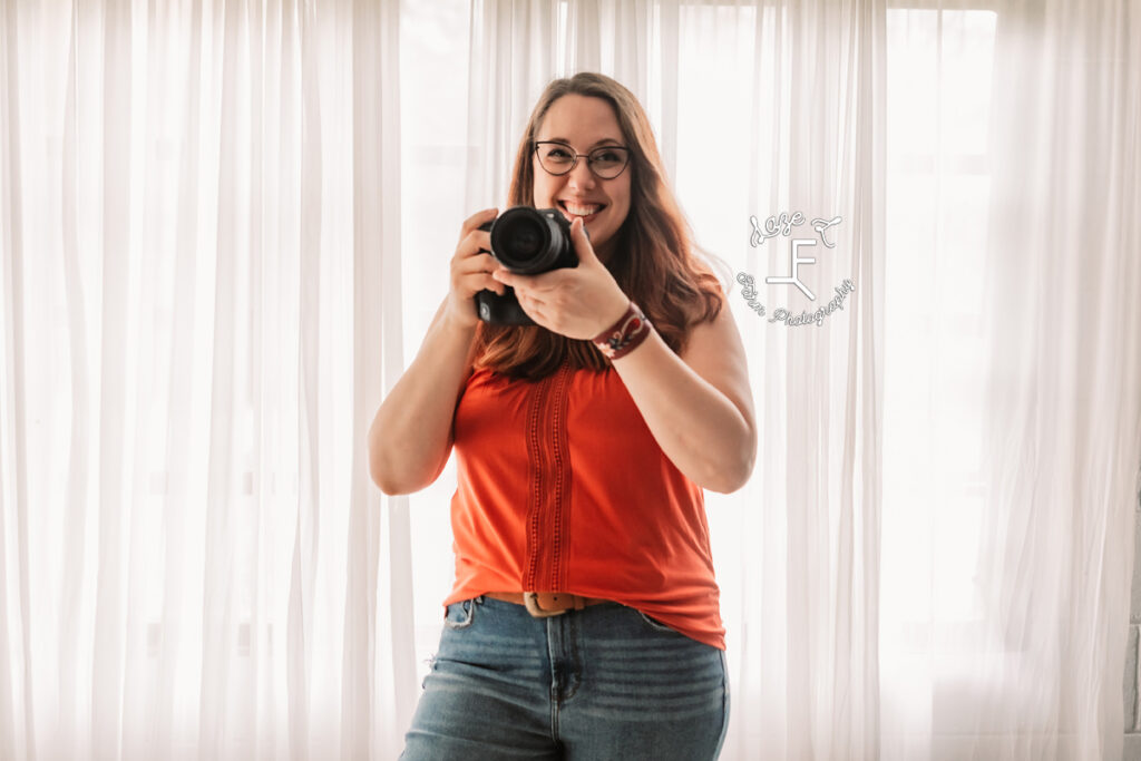 woman in front of white curtains in orange shirt holding camera