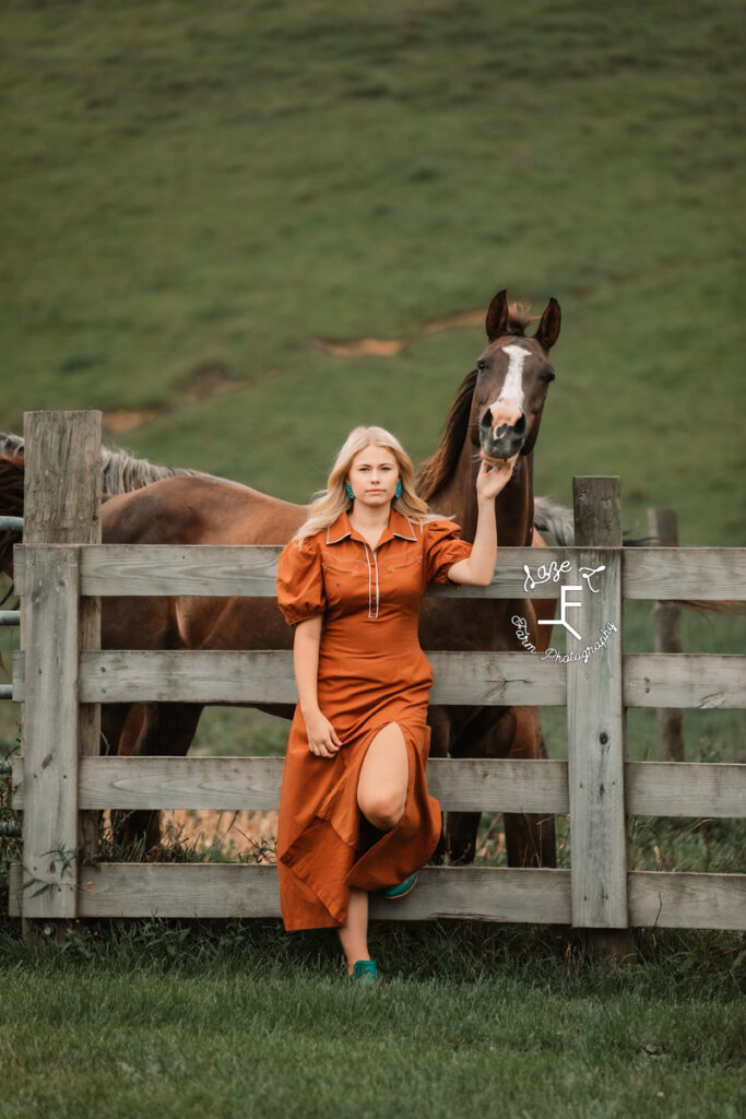 model in burnt orange dress leaning against fence petting horse