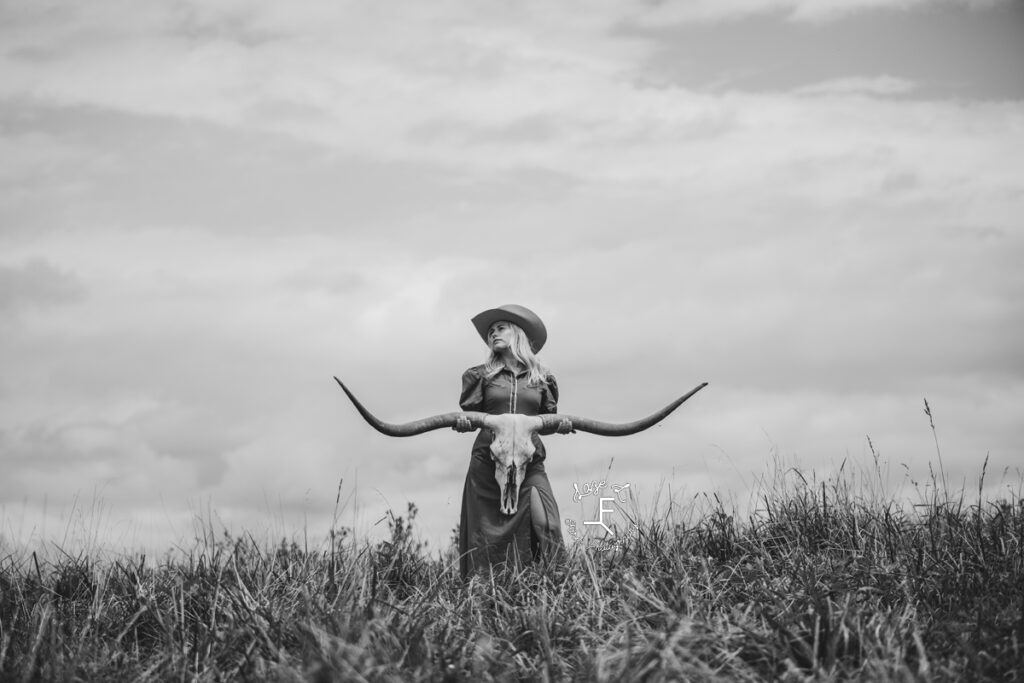cowgirl standing on top of hill holding long horn skull in black and white