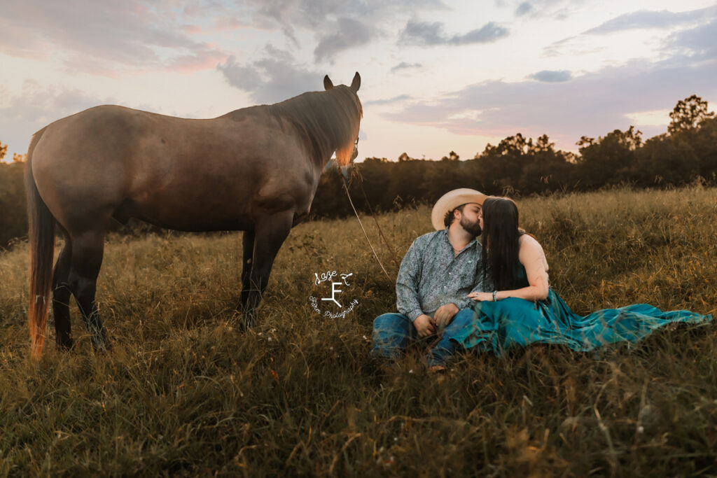 cowboy and fiancé kissing sitting in field with horse