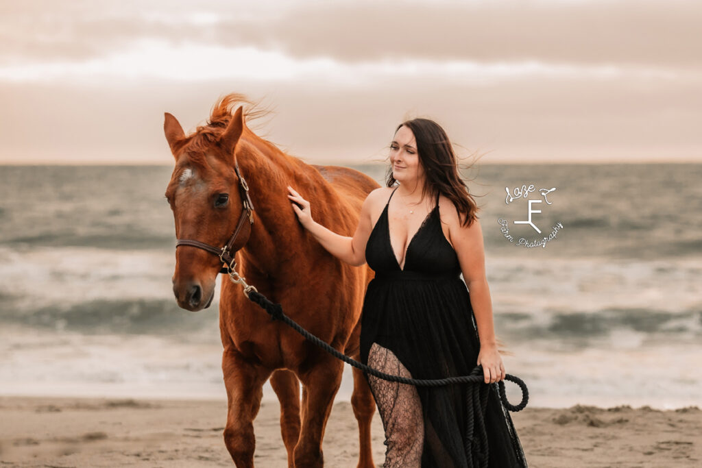 cowgirl in black dress walking on beach with older mare