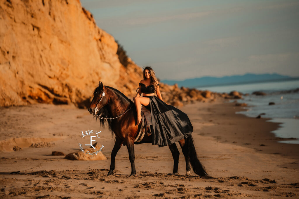 model in black dress riding bay horse on the beach