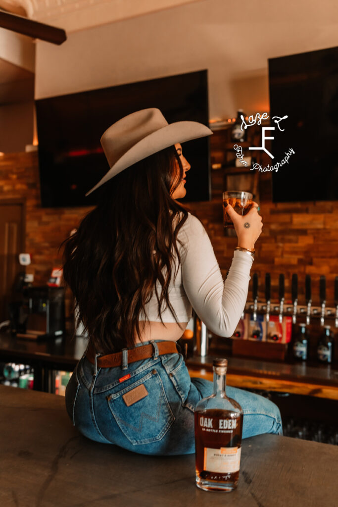 cowgirl sitting on bar with bottle of whiskey beside her