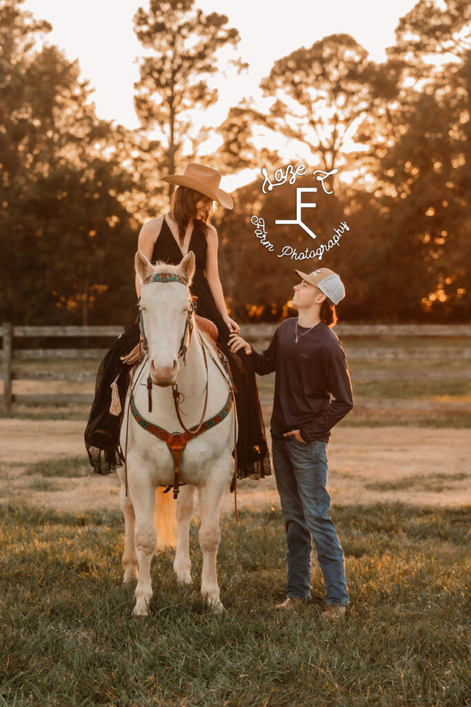 cowgirl in black dress sitting on white horse looking down at boyfriend on the ground