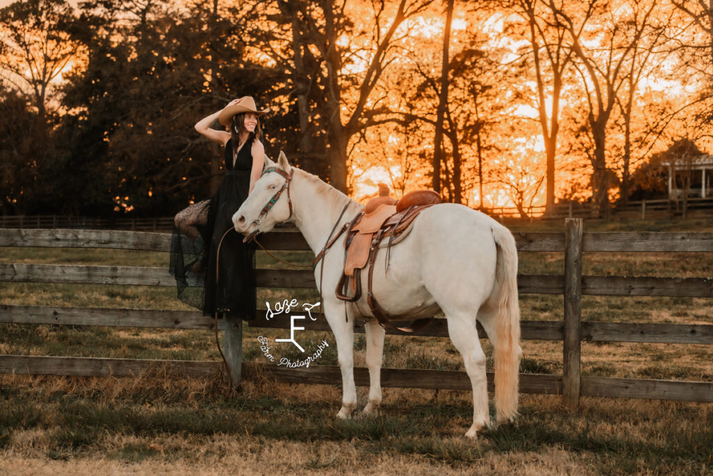 cowgirl in black dress sitting on fence at sunset with white horse