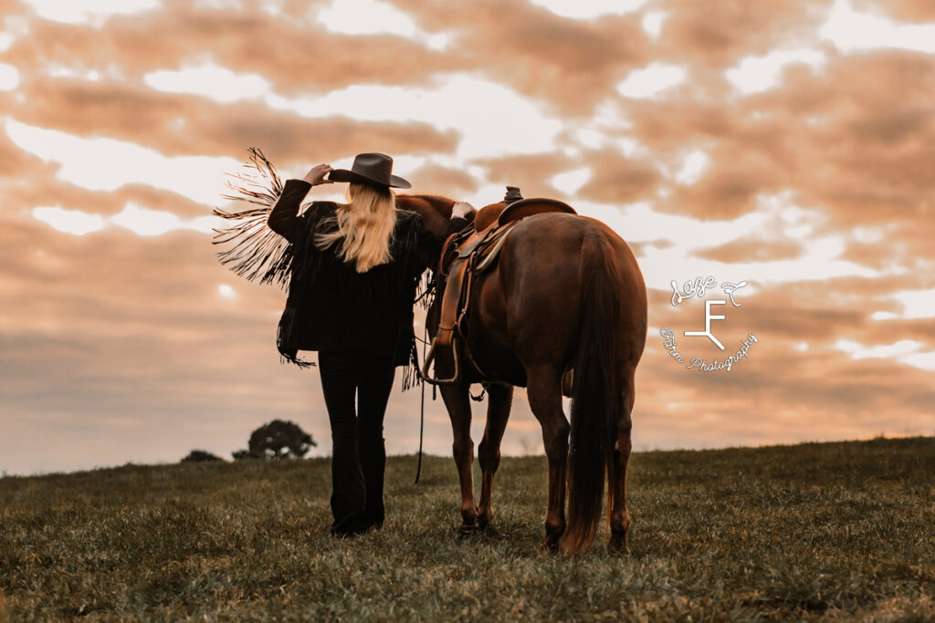 cowgirl in fringe jacket facing away from camera with her horse