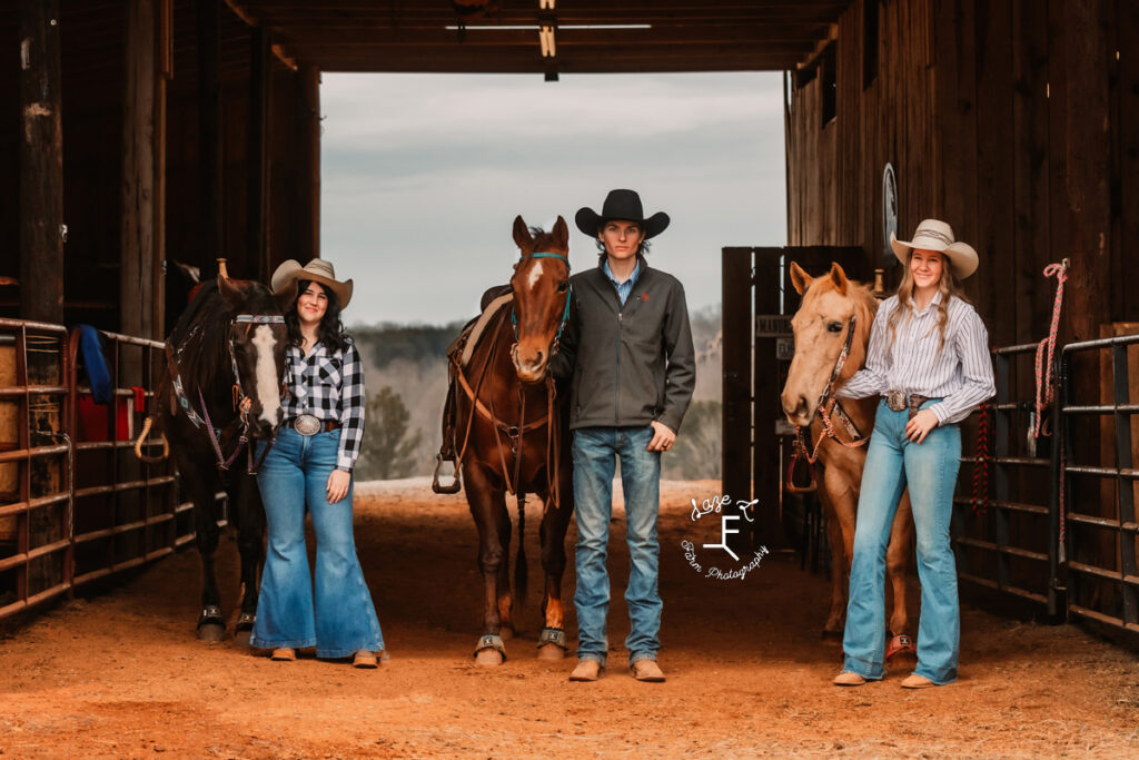 2 cowgirls and a cowboy with their horses in the barn aisle