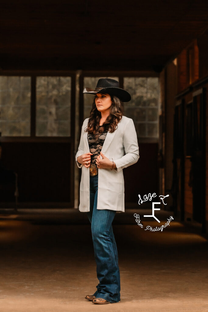 Amy standing in barn aisle in white blazer and black hat with jeans