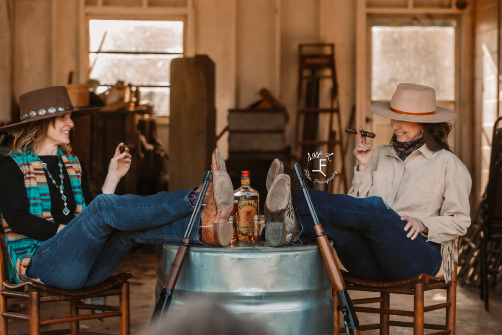 Gina and Christina sitting with feet propped up at table with whiskey and cigars