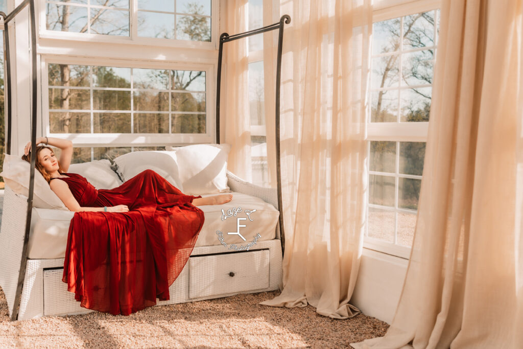 Bella in deep red dress laying on day bed looking out window