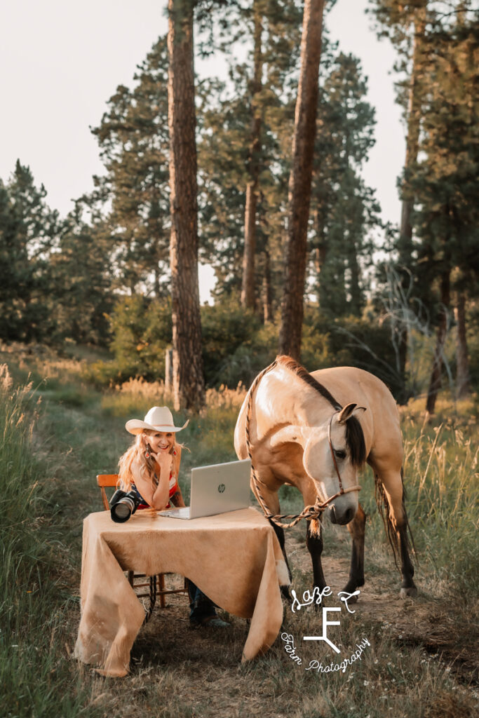 Theresa sitting at table with laptop, camera and her horse standing