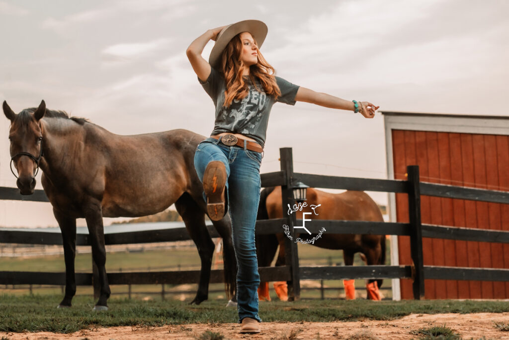 Kendall holding hat kicking boot toward camera with horses in background