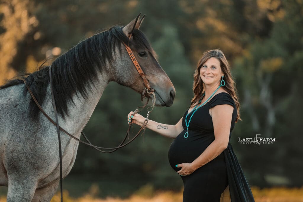 A girl standing beside  her husband and horse in a black maternity dress during a photoshoot with Laze L Farm Photography