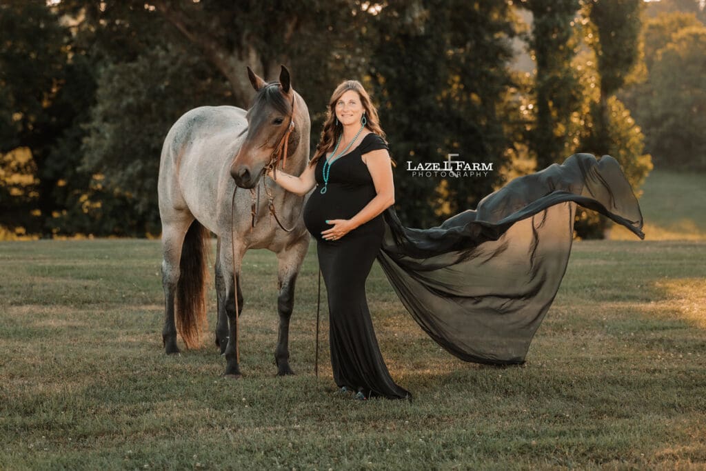 A girl standing beside her horse in a black maternity dress during a photoshoot with Laze L Farm Photography