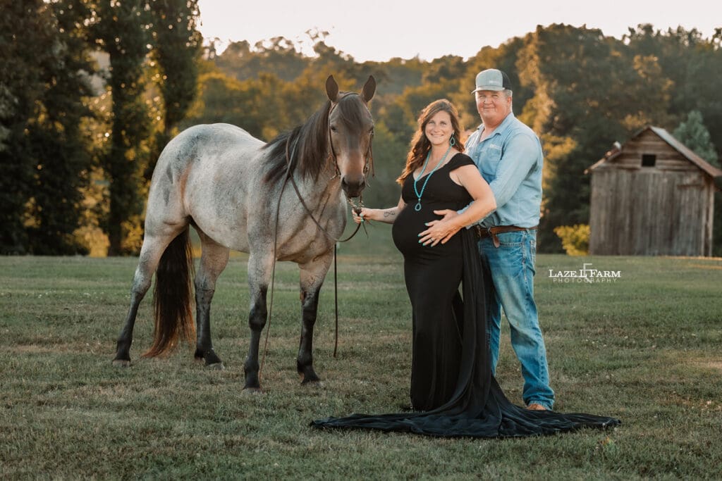 A girl standing beside  her husband and horse in a black maternity dress during a photoshoot with Laze L Farm Photography