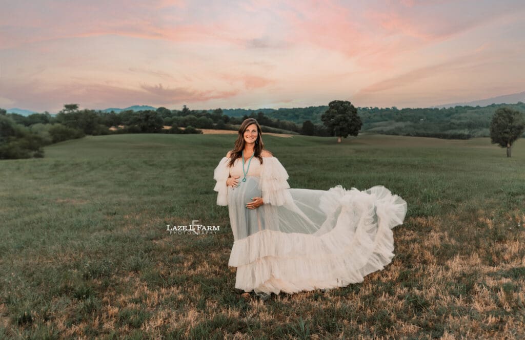 A girl standing in a middle of the field at sunset during her maternity session in a white dress during a photoshoot with Laze L Farm Photography