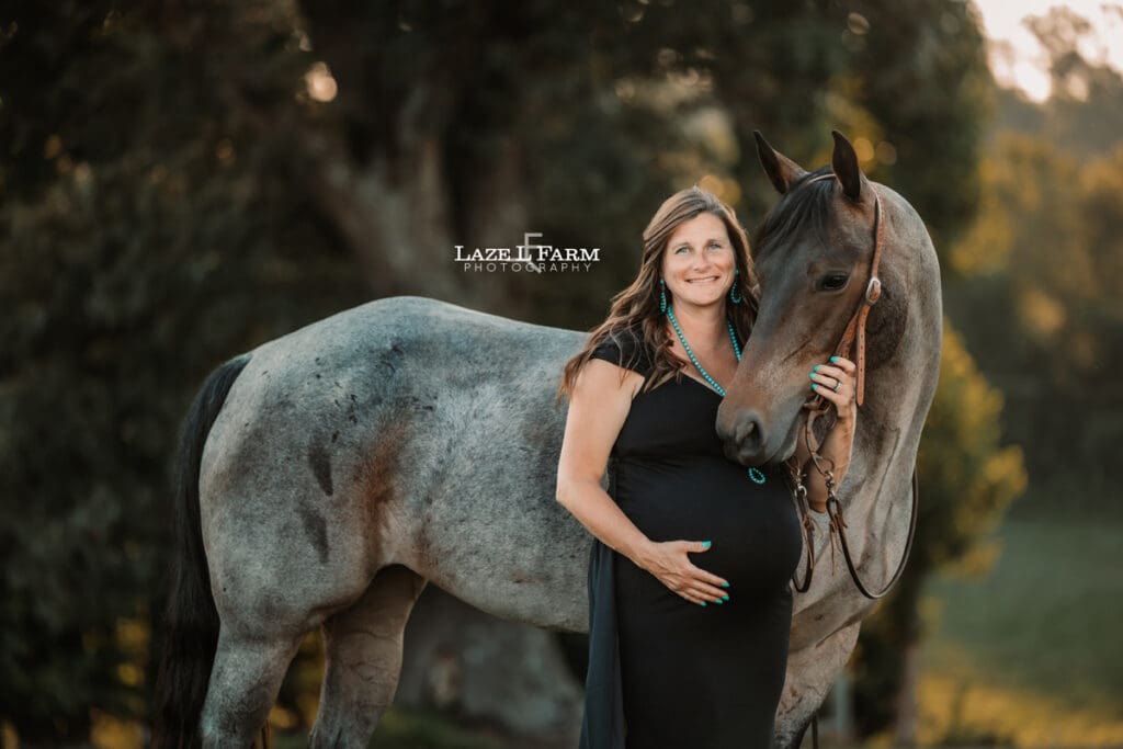 A girl standing beside  her husband and horse in a black maternity dress during a photoshoot with Laze L Farm Photography